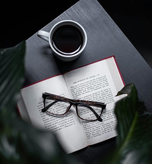 A serene setting with an open book, eyeglasses, and a steaming cup of coffee on a dark table.