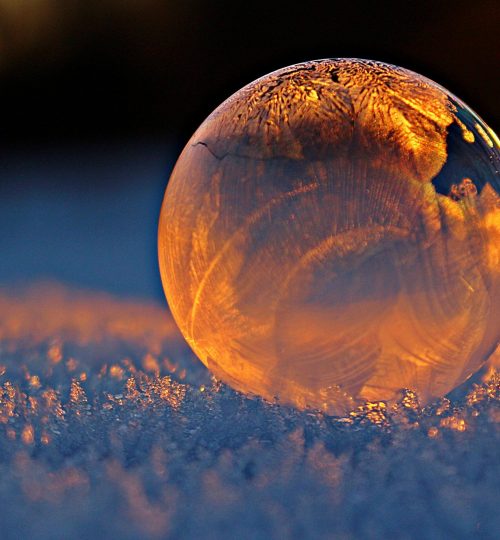Close-up shot of a frozen bubble with warm reflections resting on a snowy surface at twilight.